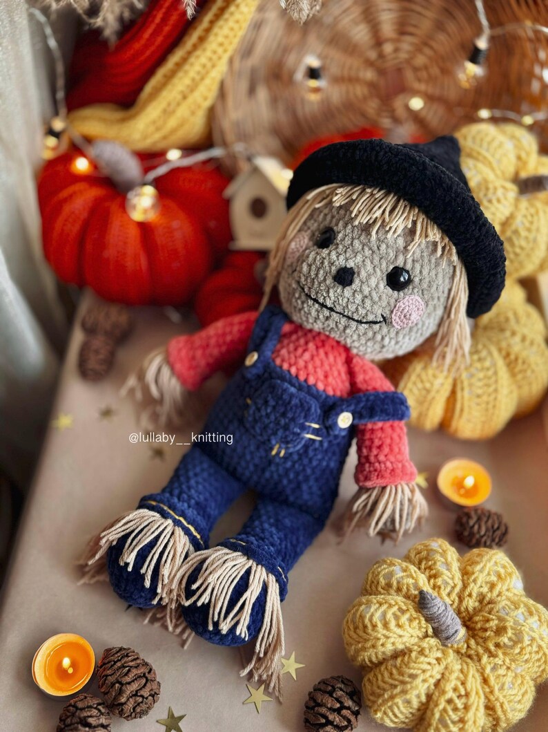 May include: A crocheted scarecrow doll wearing a black hat, red shirt, and blue overalls. The doll is surrounded by crocheted pumpkins and fall decor.