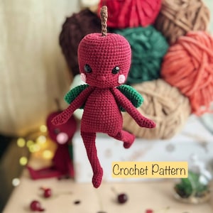Puede incluir: Una muñeca de crochet con forma de manzana con alas verdes y cuerpo rojo. La muñeca está colgada en el aire sobre una mesa con cerezas rojas esparcidas sobre la superficie. El texto "Crochet Pattern" es visible en la imagen.
