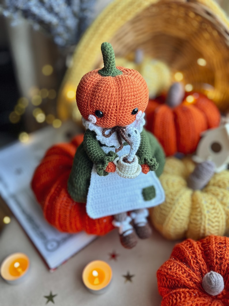 Op de afbeelding: Een gehaakte pompoendoll die een groene jurk en een witte schort draagt, met een kopje koffie in de hand. De pop is omringd door andere gehaakte pompoenen in oranje, geel en bruin.