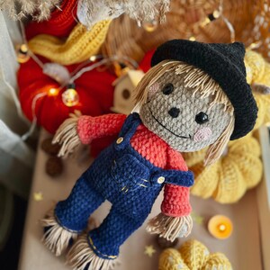 May include: A crocheted scarecrow doll wearing a black hat, red shirt, and blue overalls. The doll is surrounded by crocheted pumpkins and fall decor.