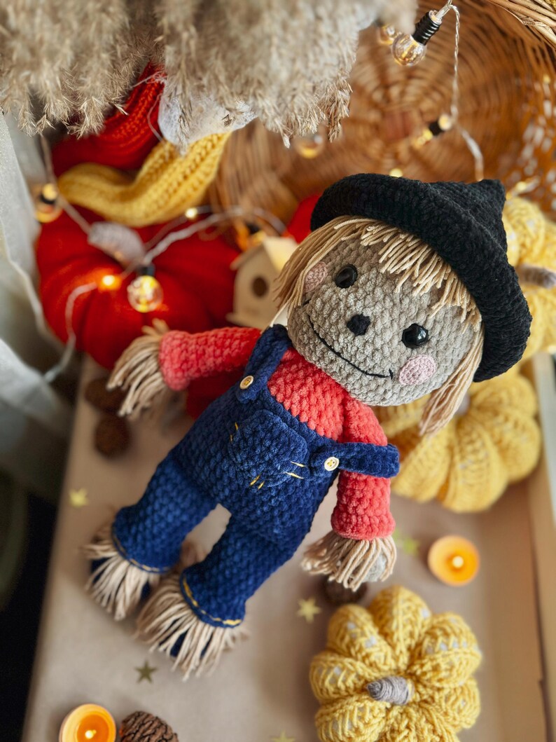 May include: A crocheted scarecrow doll wearing a black hat, red shirt, and blue overalls. The doll is surrounded by crocheted pumpkins and fall decor.