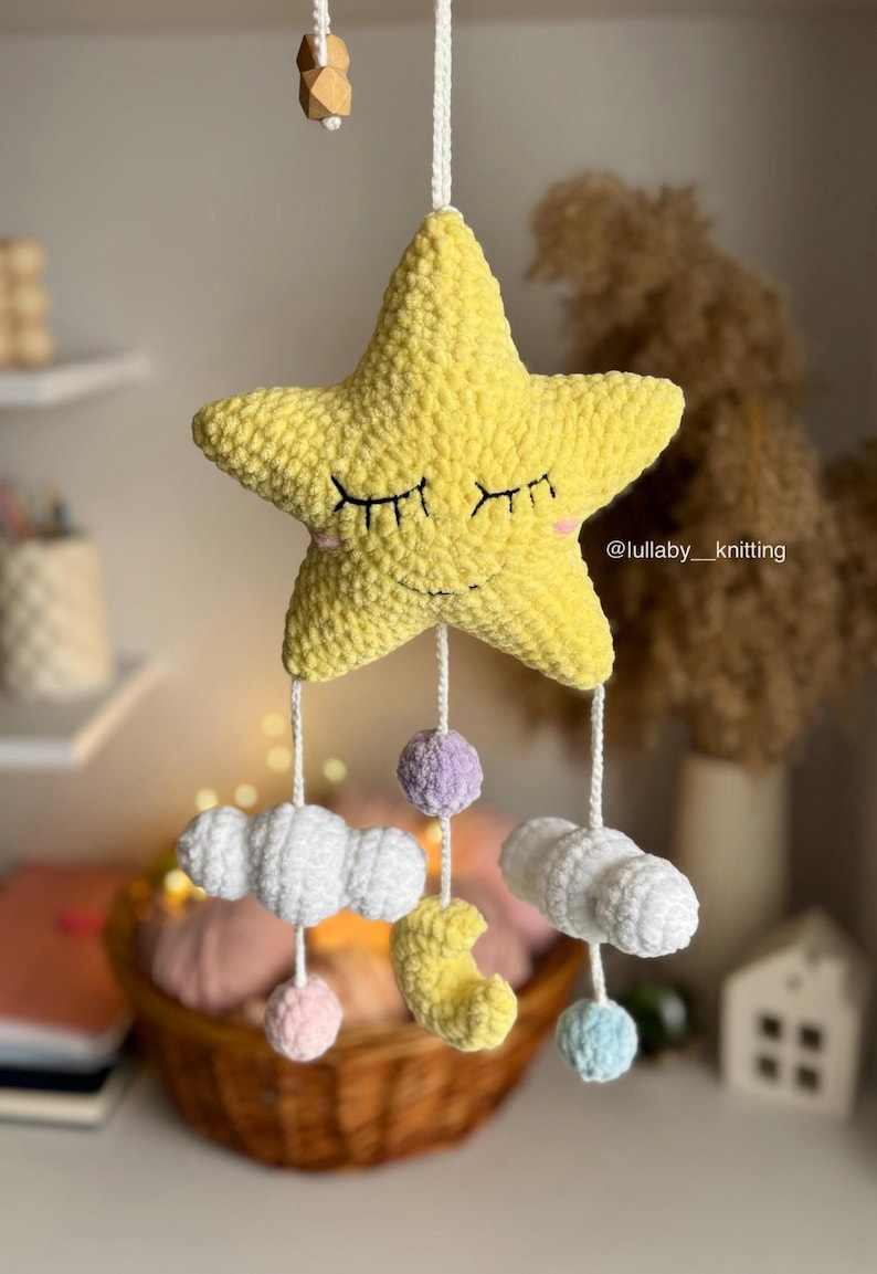 May include: A handmade yellow star mobile with closed eyes, clouds, moon, and pom-poms. The star is the focal point, with dangling elements in white, pink, purple, and blue. The mobile is suspended by a white string, creating a whimsical nursery decoration.