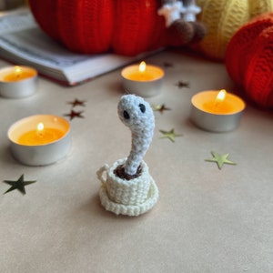 Op de afbeelding: Een wit gehaakt spookfiguurtje dat in een wit gehaakt theekopje zit. Het spook heeft twee zwarte ogen en zit op een lichtbruin oppervlak met gouden sterren eromheen.