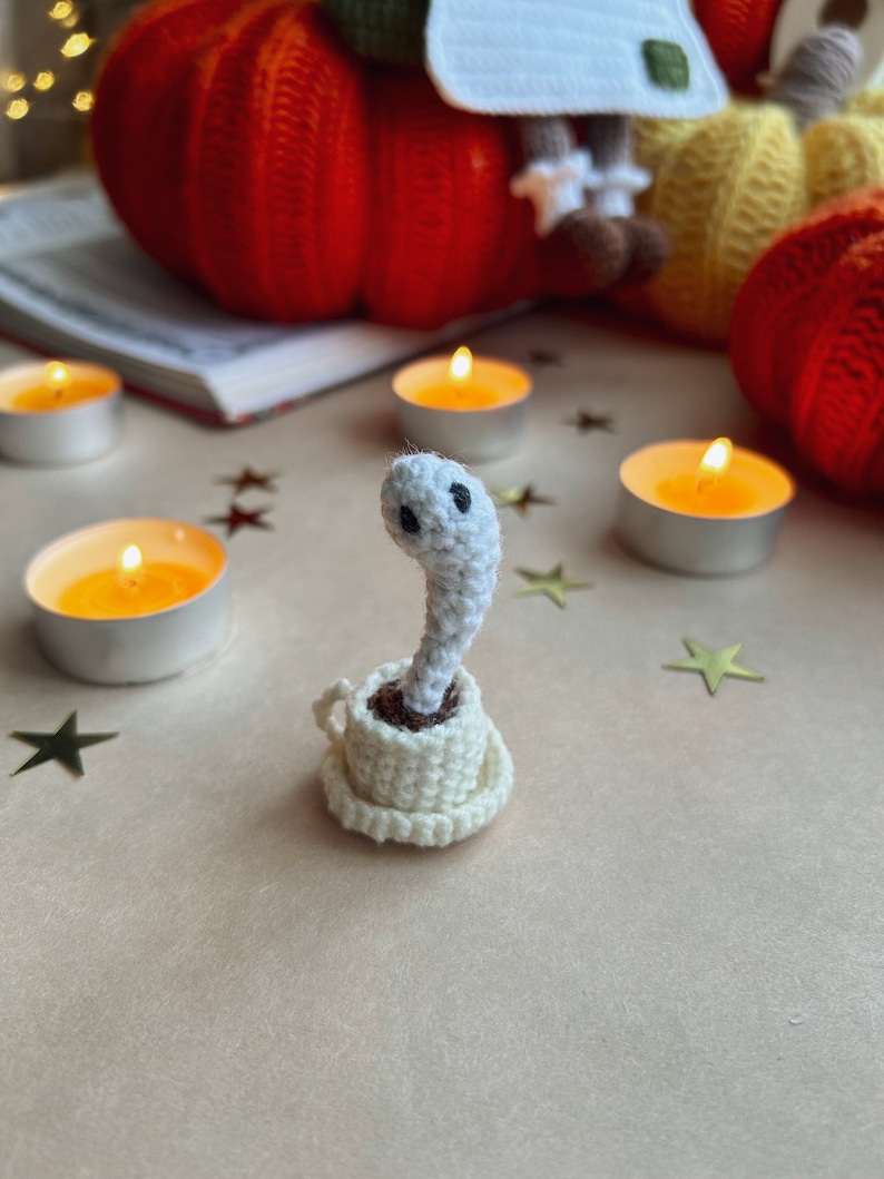 Op de afbeelding: Een wit gehaakt spookfiguurtje dat in een wit gehaakt theekopje zit. Het spook heeft twee zwarte ogen en zit op een lichtbruin oppervlak met gouden sterren eromheen.
