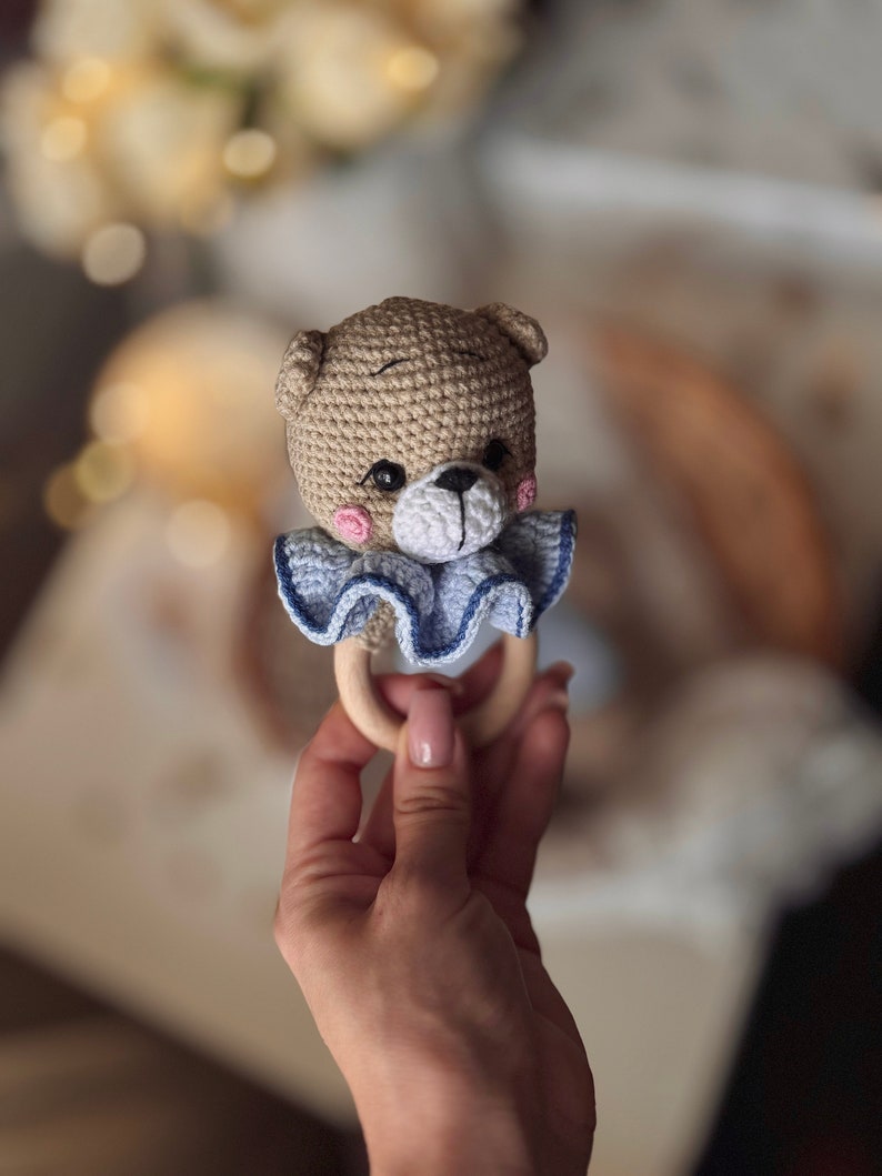 May include: A crocheted brown bear rattle with blue and white ruffle trim attached to a wooden ring.