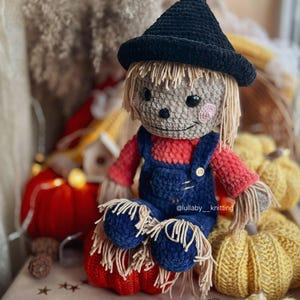 May include: A crocheted scarecrow doll wearing a black hat and blue overalls sits on a red pumpkin. The doll has a friendly face with brown eyes and a pink nose. The doll is surrounded by yellow pumpkins and other fall decorations.