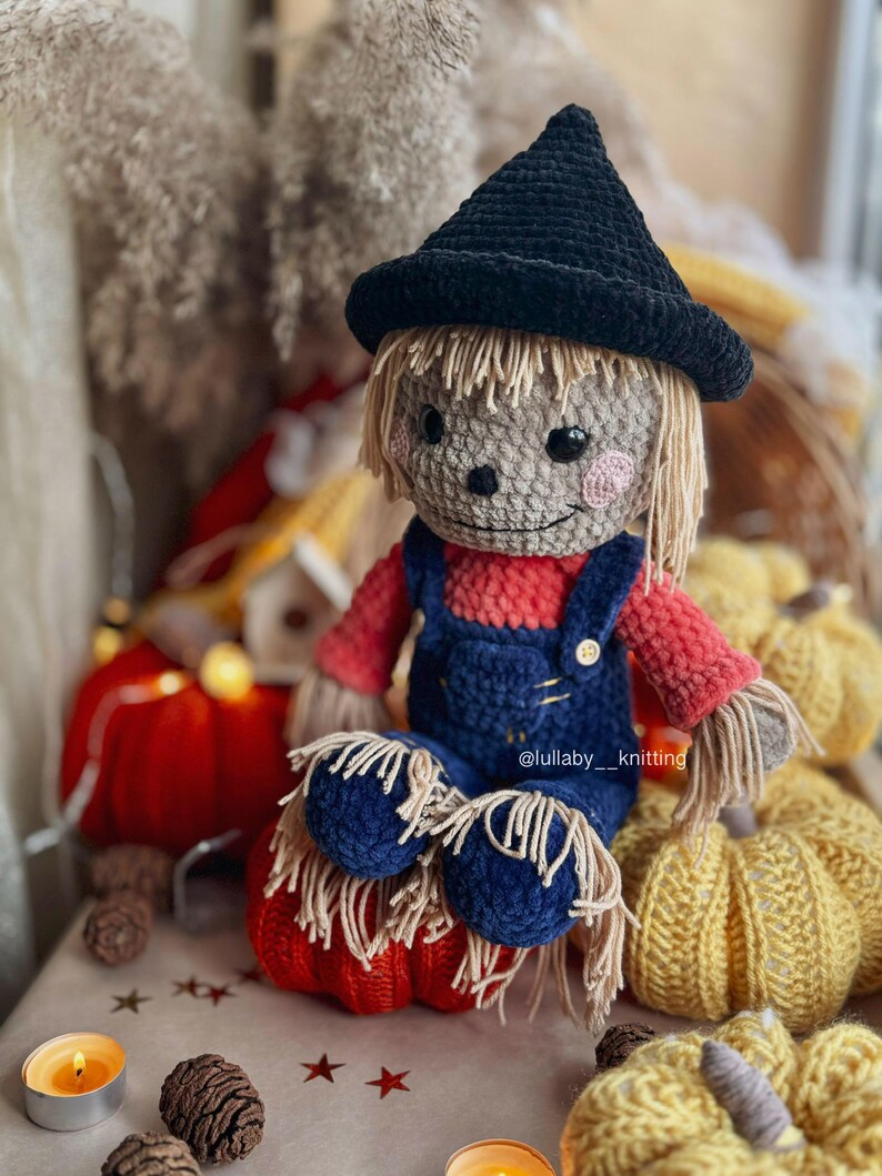 May include: A crocheted scarecrow doll wearing a black hat and blue overalls sits on a red pumpkin. The doll has a friendly face with brown eyes and a pink nose. The doll is surrounded by yellow pumpkins and other fall decorations.