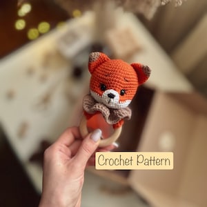 May include: A crocheted fox rattle toy with a wooden ring. The fox is orange and brown with white accents. The toy is held in a hand.