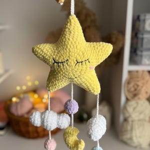 May include: A handmade, yellow crochet star mobile with closed eyes and pink cheeks. Below the star hang white clouds, a yellow crescent moon, and colorful pom-poms. The mobile is suspended by a white string with wooden beads.