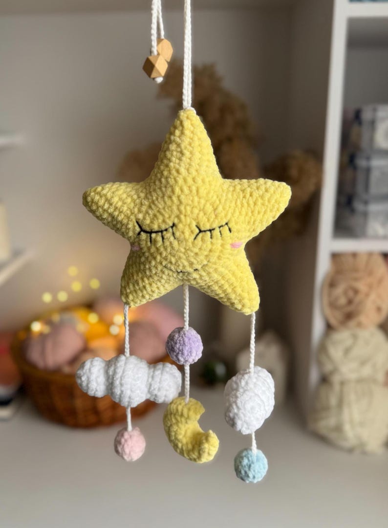 May include: A handmade, yellow crochet star mobile with closed eyes and pink cheeks. Below the star hang white clouds, a yellow crescent moon, and colorful pom-poms. The mobile is suspended by a white string with wooden beads.