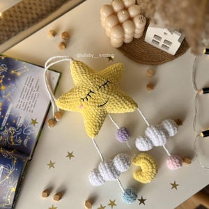 May include: A yellow crocheted star mobile with closed eyes, white clouds, a yellow crescent moon, and pastel-colored spheres. The mobile is suspended by white strings. The text "lullaby_knitting" is visible.