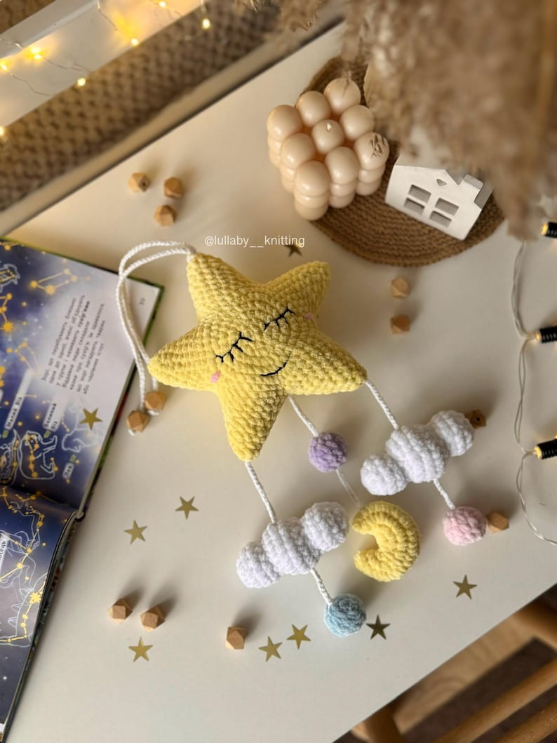 May include: A yellow crocheted star mobile with closed eyes, white clouds, a yellow crescent moon, and pastel-colored spheres. The mobile is suspended by white strings. The text "lullaby_knitting" is visible.