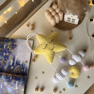 May include: A handmade yellow crochet star mobile with a smiling face, white clouds, and a yellow crescent moon. The mobile has pastel-colored accents and wooden beads. The background includes a constellation book and decorative stars.