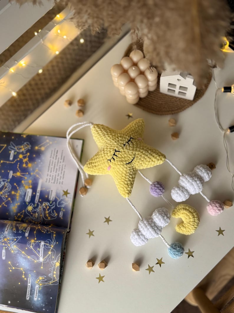 May include: A handmade yellow crochet star mobile with a smiling face, white clouds, and a yellow crescent moon. The mobile has pastel-colored accents and wooden beads. The background includes a constellation book and decorative stars.