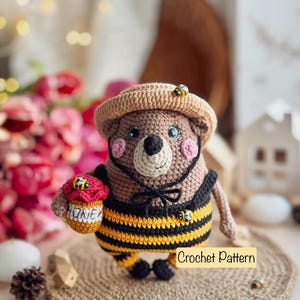 May include: Crocheted bee-themed bear wearing a brown hat and yellow and black striped outfit, holding a small jar of honey. The bear is standing on a brown surface with a "Crochet Pattern" label.