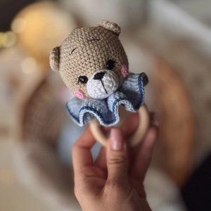 May include: A crocheted brown bear with blue and white ruffle trim attached to a wooden ring. The bear has black eyes and a black nose.