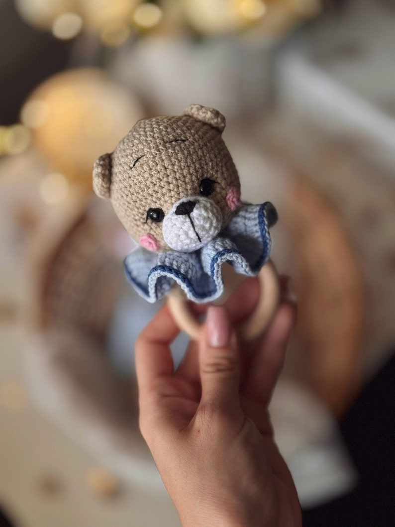 May include: A crocheted brown bear with blue and white ruffle trim attached to a wooden ring. The bear has black eyes and a black nose.