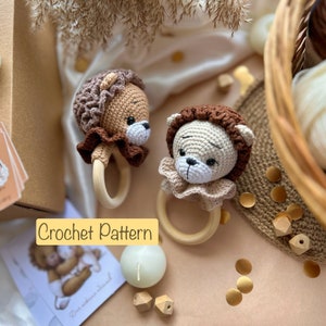 Puede incluir: Dos sonajeros de león tejidos a crochet con anillos de madera. Los leones tienen pelaje marrón y beige con ojos y narices marrones. Los juguetes están sobre una superficie marrón claro con una caja de madera y una tarjeta que dice "Crochet Pattern".