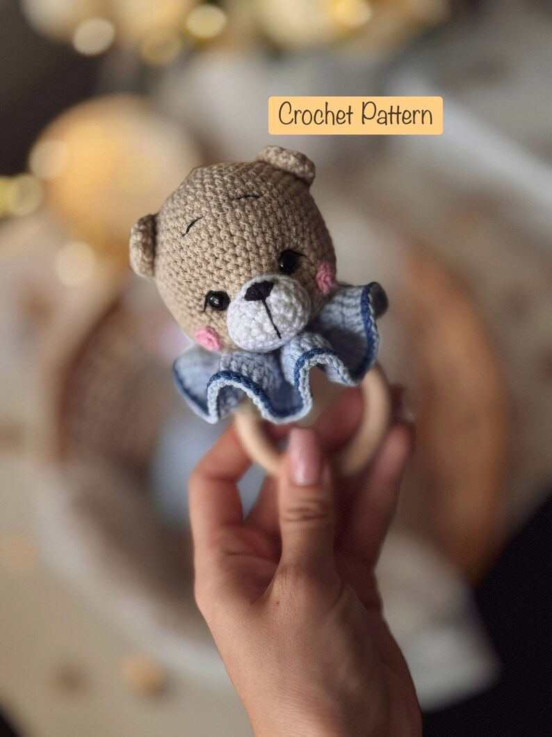 May include: Crocheted teddy bear rattle with a blue and white ruffled collar, attached to a wooden ring. Crochet Pattern.