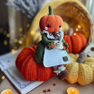 Op de afbeelding: Een gehaakte pompoendoll die een groene jurk draagt en een kopje vasthoudt. De pop zit op een grote oranje gehaakte pompoen. Er zijn twee kleinere gehaakte pompoenen op de achtergrond, een gele en een oranje.