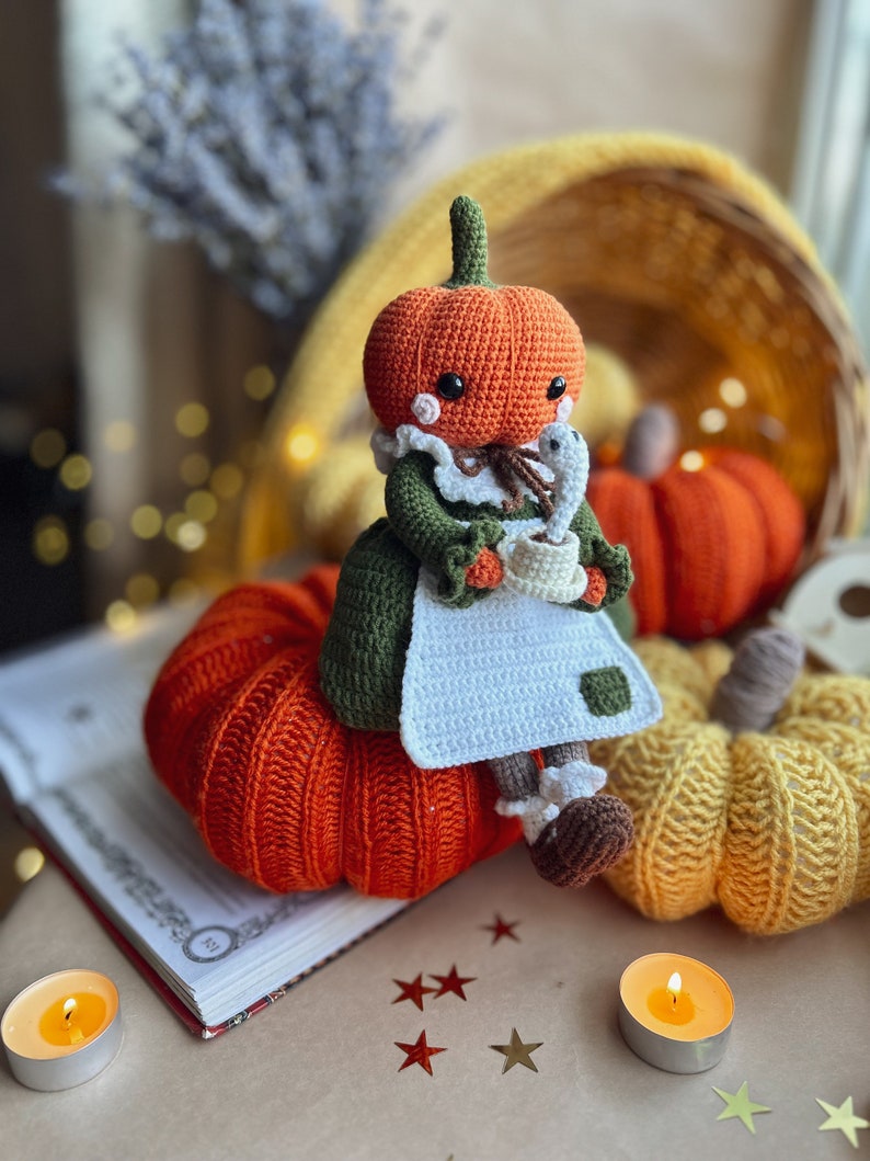 Op de afbeelding: Een gehaakte pompoendoll die een groene jurk draagt en een kopje vasthoudt. De pop zit op een grote oranje gehaakte pompoen. Er zijn twee kleinere gehaakte pompoenen op de achtergrond, een gele en een oranje.