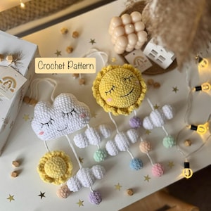 Può includere: Un mobile in crochet a forma di sole e nuvola con palline color pastello appese. Il mobile è appeso a un tavolo bianco con una superficie in legno. Il testo "Crochet Pattern" è visibile nell'immagine.