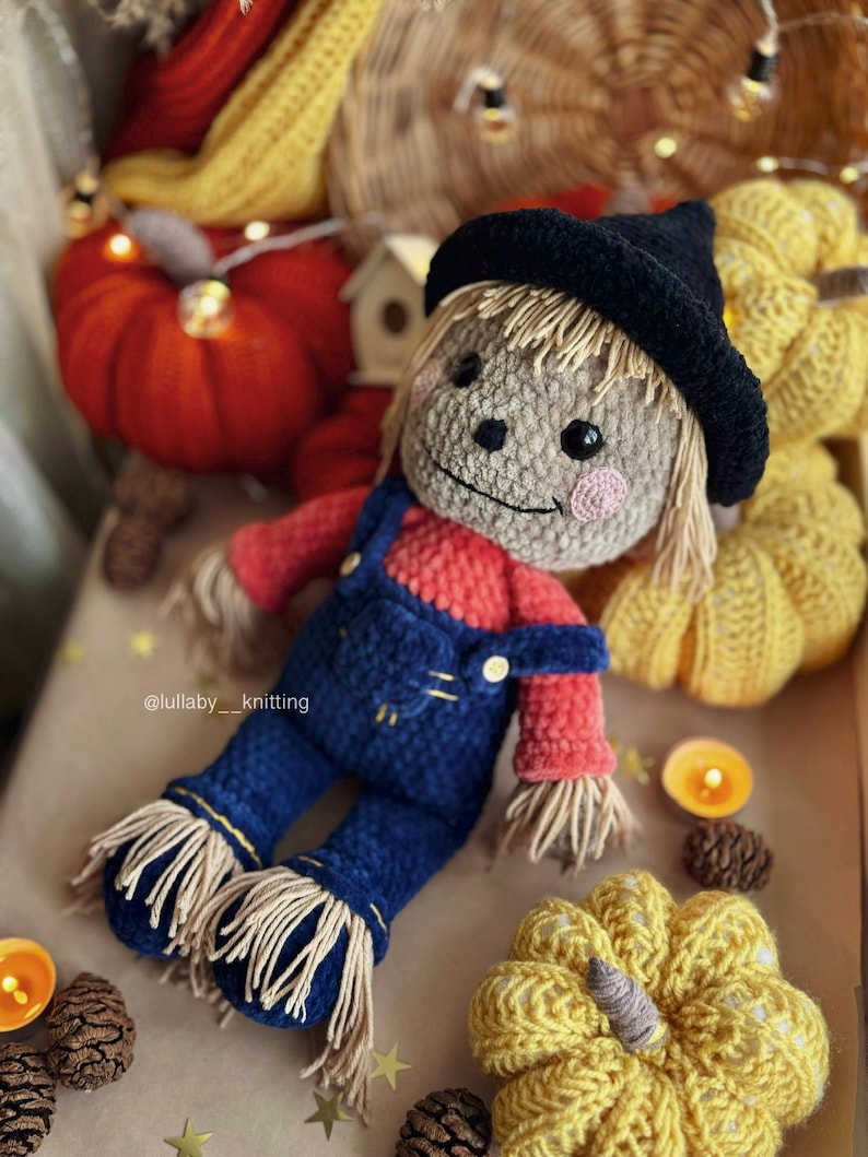May include: A crocheted scarecrow doll wearing a black hat, red and blue overalls, and a straw-like fringe. The doll is surrounded by crocheted pumpkins and other fall decorations.