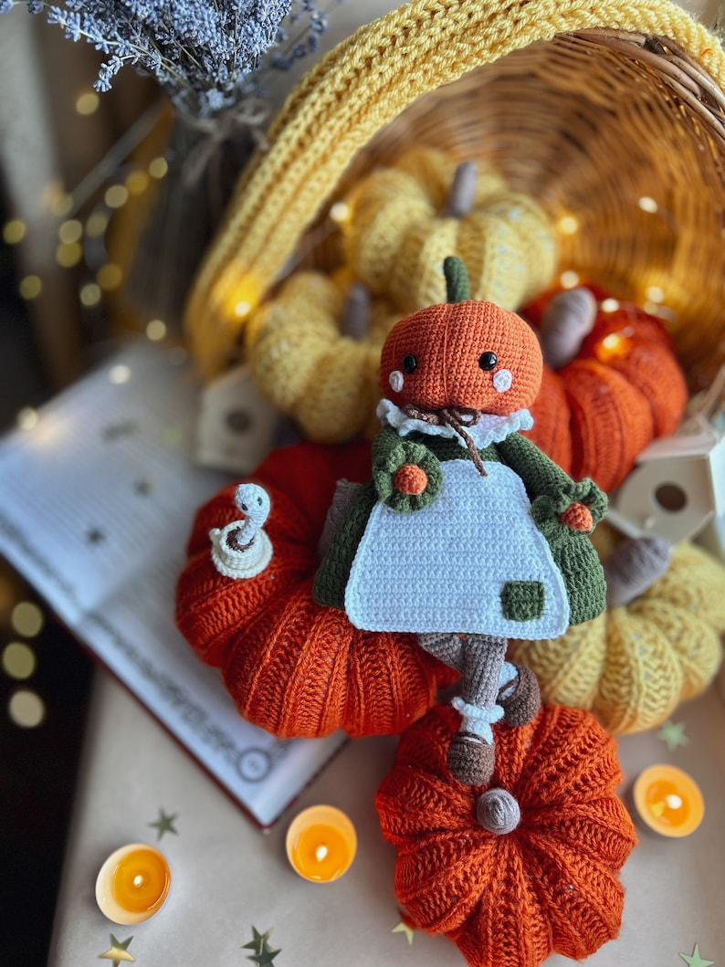 Op de afbeelding: Een gehaakte pompoendoll in een groene jurk en een witte schort zit op een boek omringd door oranje en gele gehaakte pompoenen. De sc&egrave;ne is versierd met kleine kaarsen en een rieten mand.