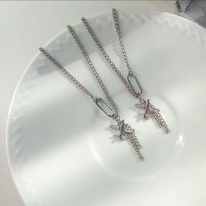 May include: Two silver-toned metal chain necklaces featuring airplane pendants. Each pendant includes a small, clear rectangular crystal set into the body of the airplane, with additional delicate chain segments hanging from the wings.