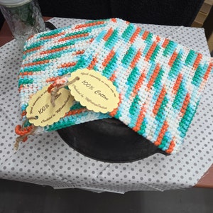 May include: Two handmade crochet potholders with a colorful striped pattern in white, teal, and orange. Each potholder has a tag that reads "100% Cotton". The potholders are resting on a dotted surface.