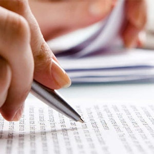May include: A person's hand holding a silver pen and writing on a white sheet of paper with black text. The paper is on a white desk with a keyboard in the background.