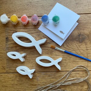 May include: A craft kit featuring three white wooden fish-shaped cutouts of varying sizes, a strip of six small paint pots in white, yellow, orange, red, blue, and green, a blue-handled paintbrush, a white card with a fish icon, and twine.