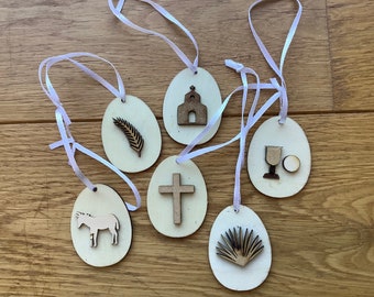 May include: Six wooden Easter egg ornaments with white ribbon hangers. Each egg features a different religious symbol: a church, a cross, a palm branch, a donkey, a chalice and a host, and a palm frond.