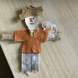 Może przedstawiać: Ręcznie robiona ozdoba w postaci stracha na wróble wykonana z drewnianej łyżki, filcu i juty. Strach na wróble ma brązowy filcowy kapelusz i koszulę, szare filcowe spodnie i ramiona z juty. Do jednego ramienia przymocowana jest naklejka z wiewiórką. Dekoracja w jesiennym klimacie.