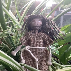 Könnte beinhalten: Eine handgefertigte Puppe mit langen braunen Haaren, die ein braunes Leinenkleid mit einem weißen Seilgürtel trägt. Die Puppe steht in einer grünen, belaubten Pflanze.