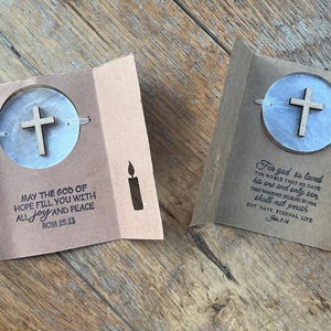 Puede incluir: Dos tarjetas plegadas abiertas con un diseño de cruz. Cada tarjeta tiene una cruz de madera en un recorte circular, con una silueta de vela y texto religioso. Las tarjetas son de papel marrón.