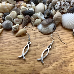 Peut inclure: Une paire de boucles d'oreilles pendantes en argent avec un charme en forme de poisson. Les boucles d'oreilles sont suspendues sur un fond de coquillages.