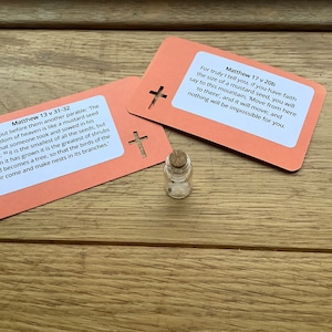 May include: Two pink cards with bible verses printed on them. The cards feature a cross cut out. A small glass bottle with a cork stopper is in the centre of the image.