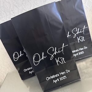 Op de afbeelding: Drie zwarte papieren zakken met witte tekst. De hoofdtekst is "Oh Shit Kit" in script. De onderkant van elke zak heeft de tekst "Christina's Hen Do April 2025". De zakken liggen op een reflecterend oppervlak.