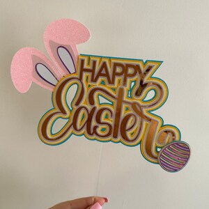 May include: A cake topper featuring pink bunny ears and the words "Happy Easter" in gold, yellow and blue lettering. A purple and gold striped Easter egg is on the right side of the topper.