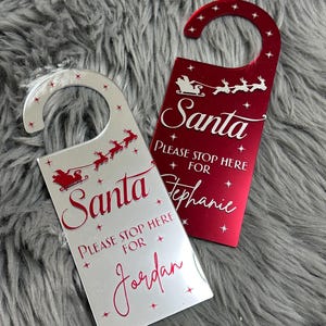 Op de afbeelding: Twee reflecterende deurhangers, één zilver en één rood, met een silhouet van de Kerstman in een slee met rendieren. De tekst luidt Santa PLEASE STOP HERE FOR Jordan en Santa PLEASE STOP HERE FOR Stephanie, versierd met kleine sterretjes.