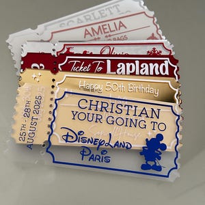 May include: A stack of decorative ticket-shaped signs. The signs have various colors, including red, gold, and white. Text on the signs includes "Ticket To Lapland", "Happy 50th Birthday", and "Christian Your Going To Disneyland Paris". A Mickey Mouse silhouette is visible.