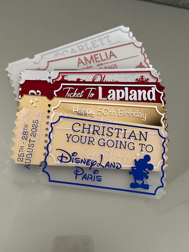 May include: A stack of decorative ticket-shaped signs. The signs have various colors, including red, gold, and white. Text on the signs includes "Ticket To Lapland", "Happy 50th Birthday", and "Christian Your Going To Disneyland Paris". A Mickey Mouse silhouette is visible.