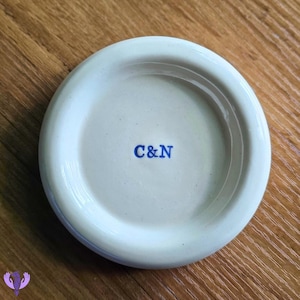 Peut inclure: Un dessous de verre rond en céramique blanc cassé avec un centre en creux. Le dessous de verre porte les lettres bleues "C&N" au centre. Il est posé sur une surface en bois.