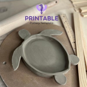 May include: A gray clay turtle-shaped dish with a raised rim. The dish is on a brown circular surface. The text "PRINTABLE -Pottery Template- " is visible above the dish.
