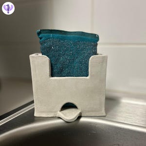 May include: A white ceramic sponge holder with a teal sponge inside. The holder has a small opening on the bottom for drainage.