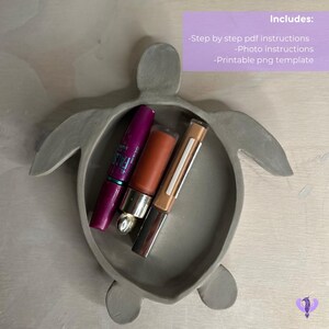 May include: A gray ceramic turtle-shaped trinket dish with three makeup items inside: a purple mascara, a brown lip gloss, and a beige lip liner.