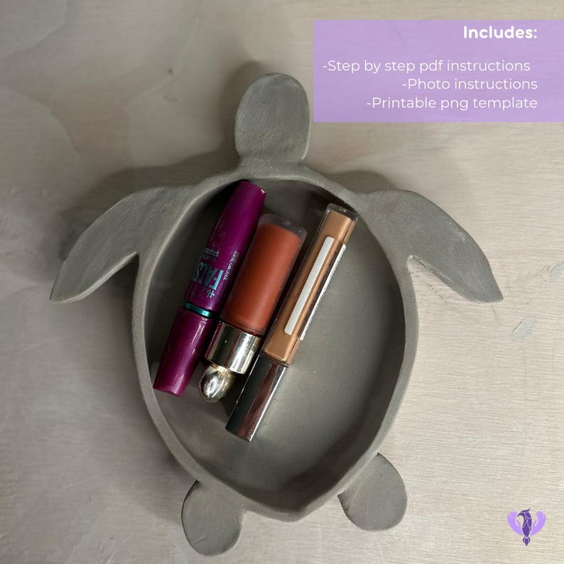 May include: A gray ceramic turtle-shaped trinket dish with three makeup items inside: a purple mascara, a brown lip gloss, and a beige lip liner.