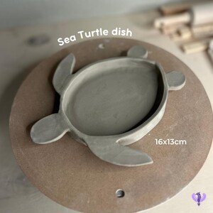May include: A gray ceramic sea turtle dish, 16 inches by 13 inches, on a brown surface.  Sea Turtle dish.
