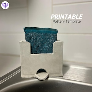 Peut inclure: Un porte-éponge en céramique blanche avec une découpe pour une éponge. Le porte-éponge a un petit bec verseur au fond pour drainer l'eau. Le porte-éponge est posé sur un évier en acier inoxydable. Le texte "PRINTABLE Pottery Template" est visible au-dessus du porte-éponge.